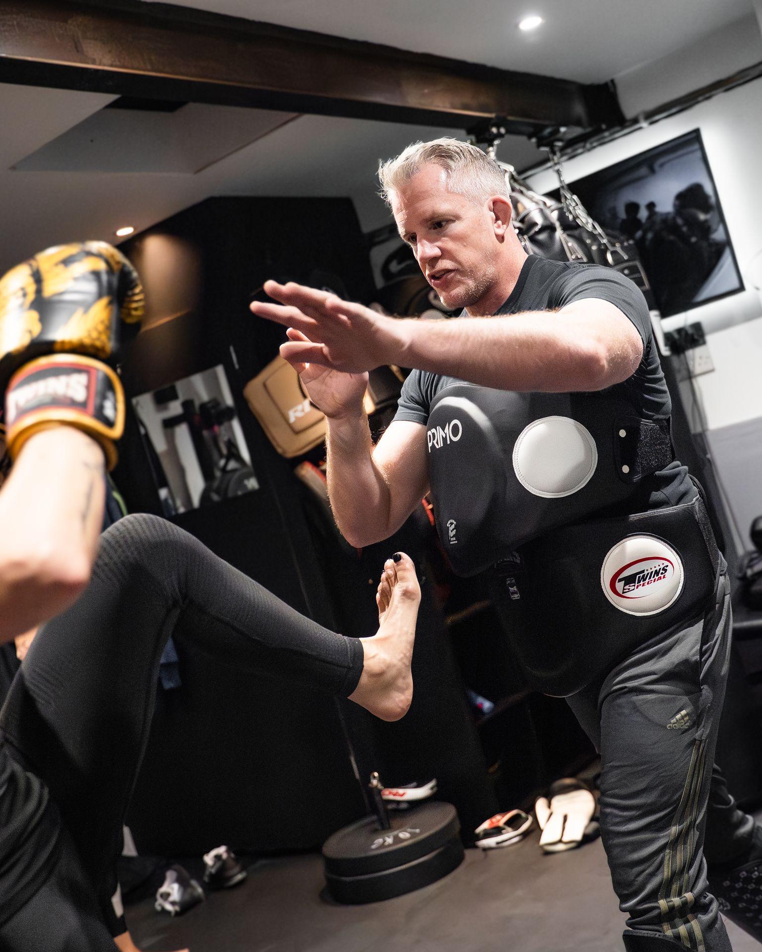 Martial arts instructor wearing protective gear coaching a student performing a kick in a gym.